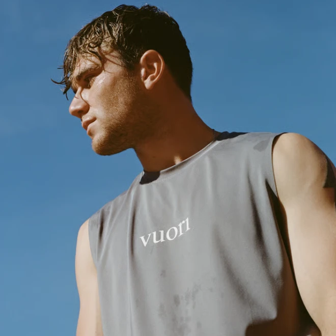 Man with wet hair wearing sleeveless gray shirt with Vuori logo against clear blue sky.