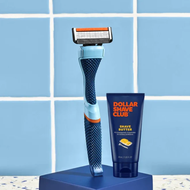 Blue and orange razor with Dollar Shave Club shave butter cream on a blue stand against blue tile.