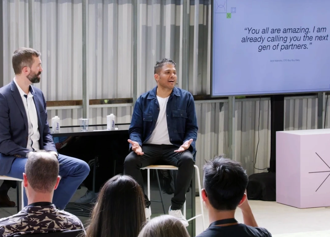 Two men seated on stage speaking to an audience with a screen displaying a motivational quote.