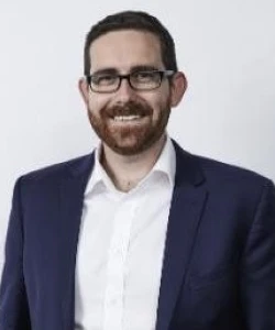 Ben Smith COO of AG1, smiling with glasses, beard, wearing a navy suit jacket and white shirt against a white background.